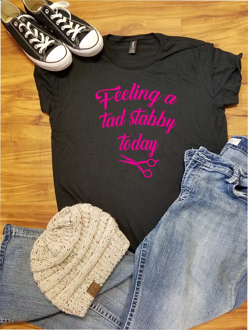 Op de afbeelding: Een casual ensemble met een zwart t-shirt met de roze tekst "Feeling a tad stabby today", gecombineerd met een blauwe jeans, een beige gebreide muts en zwarte canvas sneakers met witte veters.