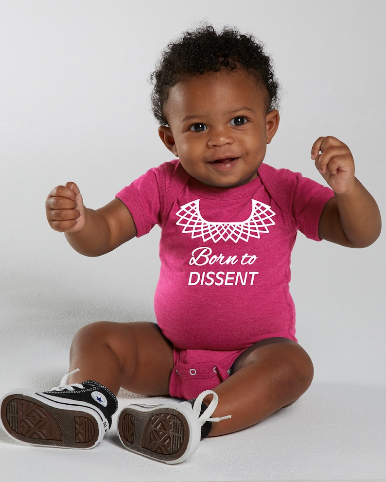 以下が含まれることがあります： 白い襟と「Born to Dissent」という文字が書かれたピンクのベビーロンパースです。