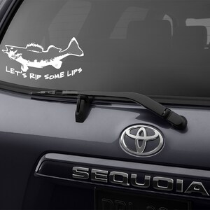 K&ouml;nnte beinhalten: Ein wei&szlig;er Vinyl-Aufkleber mit einer Silhouette eines Fisches und dem Text "Let's Rip Some Lips" auf einem grauen Auto.
