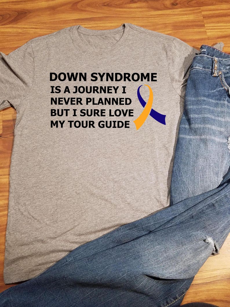 Op de afbeelding: Grijs T-shirt met een zwarte afbeelding met de tekst "Down Syndrome is a journey I never planned but I sure love my tour guide". De afbeelding bevat ook een geel en blauw lint.