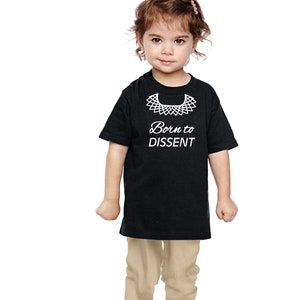 以下が含まれることがあります： 白い襟と「Born to Dissent」という文字の白いグラフィックがプリントされた黒いTシャツ。小さな子供がTシャツを着ています。