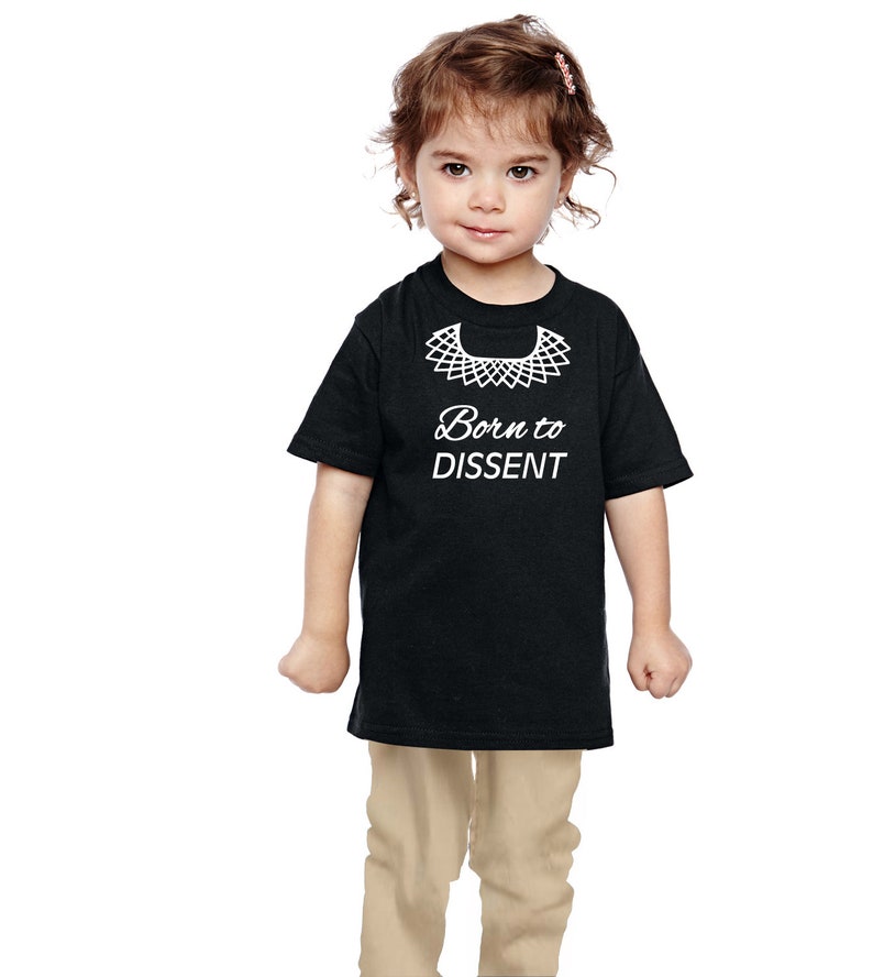 以下が含まれることがあります： 白い襟と「Born to Dissent」という文字の白いグラフィックがプリントされた黒いTシャツ。小さな子供がTシャツを着ています。