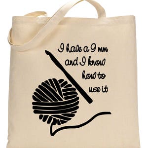May include: A beige canvas tote bag with black text and a graphic of a ball of yarn and a crochet hook. The text reads "I have a 9 mm and I know how to use it."