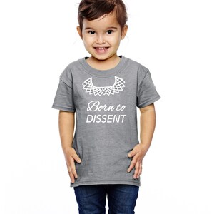 以下が含まれることがあります： 白い襟と「Born to Dissent」という文字が書かれたグレーのTシャツを着た幼い子供。子供は笑顔で、髪はポニーテールに結ばれています。
