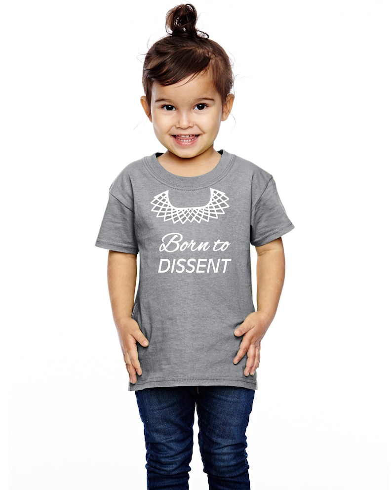 以下が含まれることがあります： 白い襟と「Born to Dissent」という文字が書かれたグレーのTシャツを着た幼い子供。子供は笑顔で、髪はポニーテールに結ばれています。
