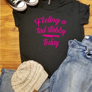Op de afbeelding: Zwart T-shirt met de roze tekst "Feeling a tad stabby today", een roze mesafbeelding en een paar zwarte canvas sneakers. Een lichtbeige gebreide muts en een blauwe jeans maken de casual outfit compleet.