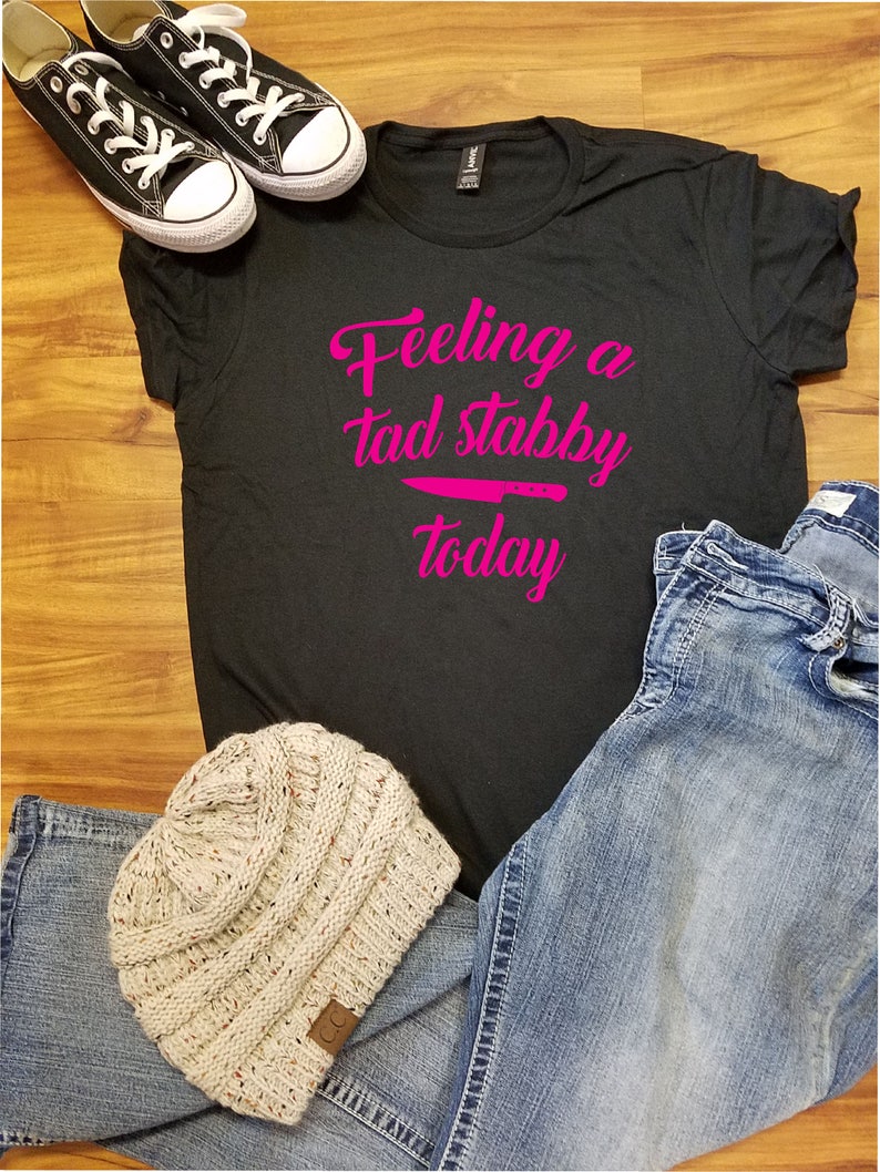 Op de afbeelding: Zwart T-shirt met de roze tekst "Feeling a tad stabby today", een roze mesafbeelding en een paar zwarte canvas sneakers. Een lichtbeige gebreide muts en een blauwe jeans maken de casual outfit compleet.
