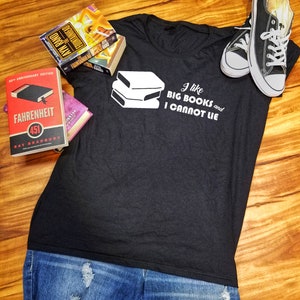 May include: A black t-shirt with a white graphic of stacked books and the text "I like BIG BOOKS and I CANNOT LIE". The shirt is laying on a wooden surface with a pair of black sneakers and three books.