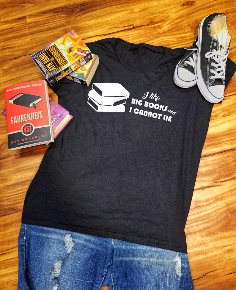 May include: A black t-shirt with a white graphic of stacked books and the text "I like BIG BOOKS and I CANNOT LIE". The shirt is laying on a wooden surface with a pair of black sneakers and three books.