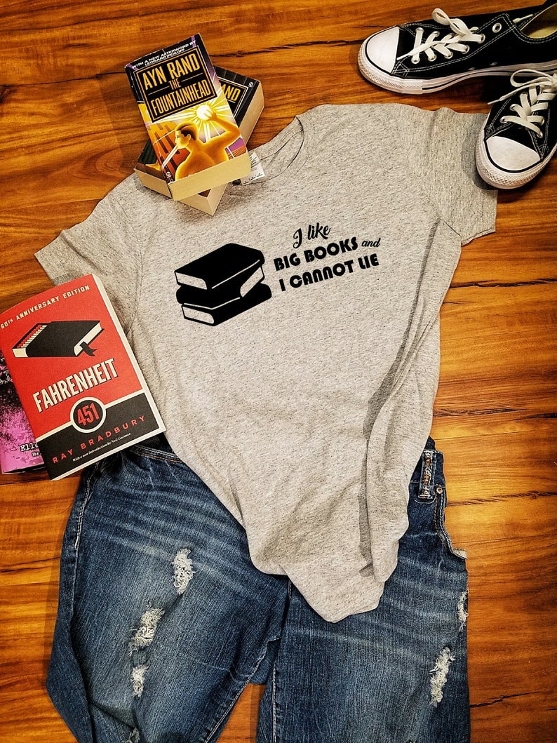 May include: A gray t-shirt with the text "I like big books and I cannot lie" printed on it. The shirt is laying on a wooden surface with two books and a pair of jeans.