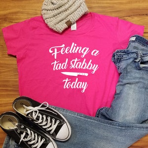 Op de afbeelding: Een casual outfit met een felroze t-shirt met de tekst "Feeling a tad stabby today" en een mesafbeelding. Inclusief een grijze beanie, een blauwe jeans en zwarte Converse sneakers met witte veters.