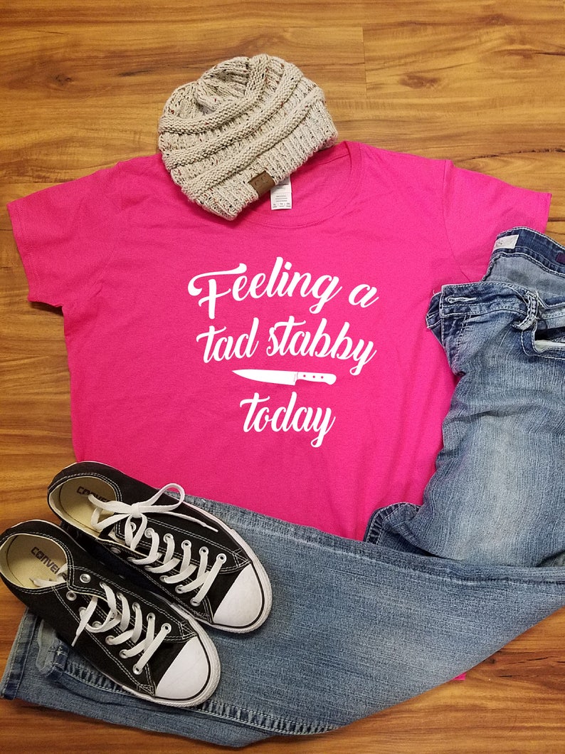 Op de afbeelding: Een casual outfit met een felroze t-shirt met de tekst "Feeling a tad stabby today" en een mesafbeelding. Inclusief een grijze beanie, een blauwe jeans en zwarte Converse sneakers met witte veters.