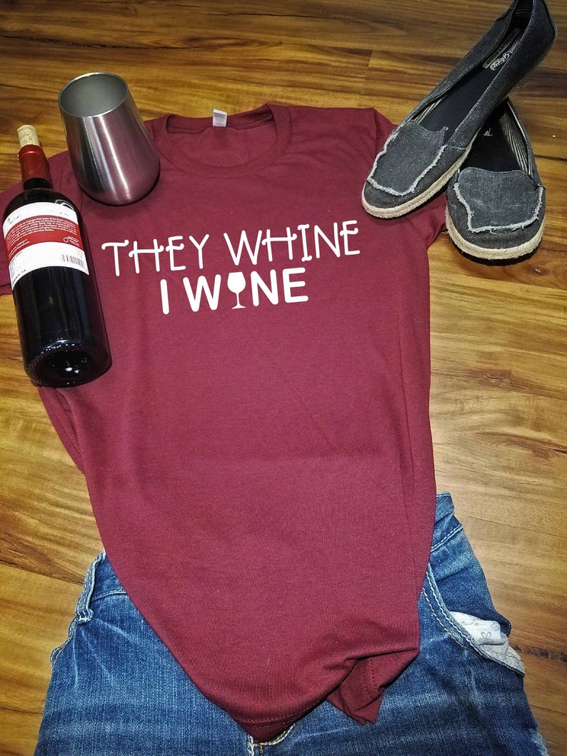 K&ouml;nnte beinhalten: Ein maroonfarbenes T-Shirt mit dem Text "They whine I wine" und einer Grafik eines Weinglases. Das Shirt liegt auf einer blauen Jeans und einer Holzoberfl&auml;che. Auf der Oberfl&auml;che befinden sich eine Weinflasche und ein silbernes Weinglas.