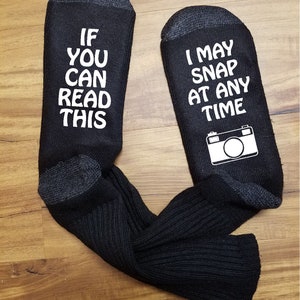 May include: A pair of black socks with white text. One sock says "If you can read this" and the other sock says "I may snap at any time" with a camera icon.