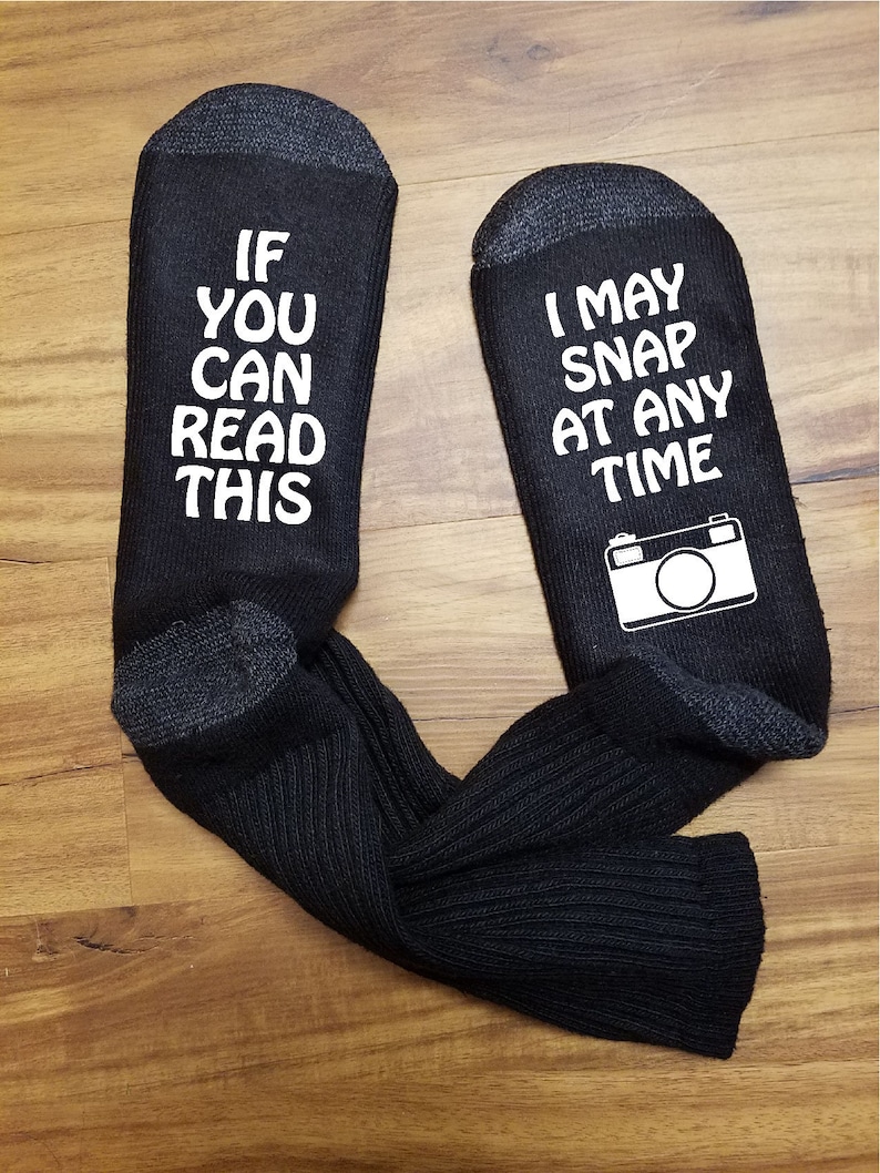 May include: A pair of black socks with white text. One sock says "If you can read this" and the other sock says "I may snap at any time" with a camera icon.