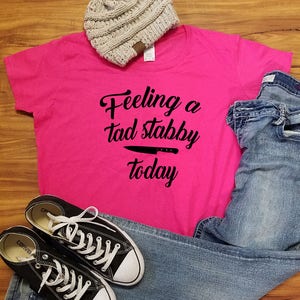 Op de afbeelding: Een felroze t-shirt met de tekst "Feeling a tad stabby today" en een mesafbeelding. De afbeelding bevat ook een grijze muts, een blauwe jeans en zwarte Converse sneakers met witte veters.