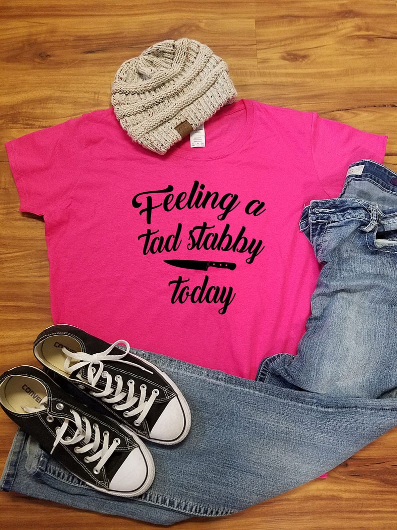 Op de afbeelding: Een felroze t-shirt met de tekst "Feeling a tad stabby today" en een mesafbeelding. De afbeelding bevat ook een grijze muts, een blauwe jeans en zwarte Converse sneakers met witte veters.
