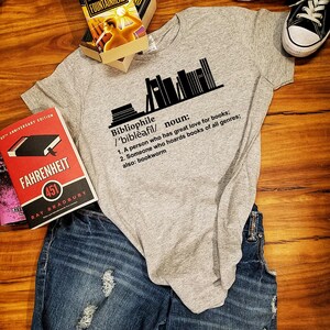 May include: A gray t-shirt with a black graphic of a stack of books and the definition of the word "bibliophile". The text reads: "Bibliophile /'bibleə,fil/ noun: 1. A person who has great love for books; 2. Someone who hoards books of all genres; also: bookworm"