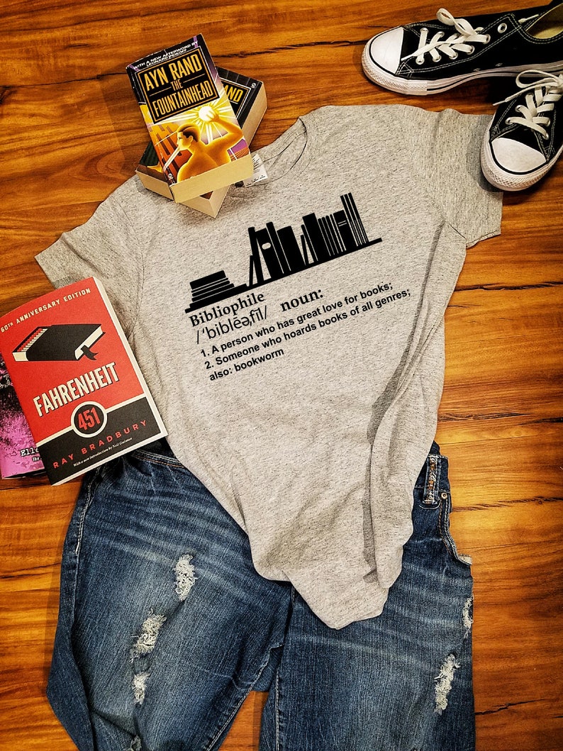May include: A gray t-shirt with a black graphic of a stack of books and the definition of the word "bibliophile". The text reads: "Bibliophile /'bibleə,fil/ noun: 1. A person who has great love for books; 2. Someone who hoards books of all genres; also: bookworm"