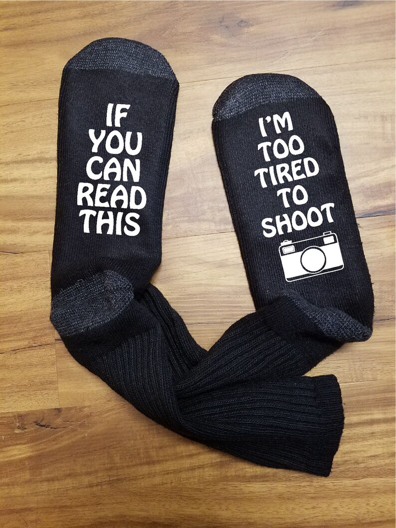 May include: A pair of black socks with white text. One sock reads "If you can read this" and the other sock reads "I'm too tired to shoot" with a camera icon.
