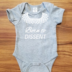 以下が含まれることがあります： 白い襟のデザインと「Born to Dissent」というテキストが書かれた灰色のベビーロンパースです。