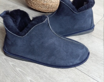 May include: A pair of navy blue slippers with a soft, dark interior lining. The exterior features a smooth, matte finish with visible stitching along the seams and a small pull tab at the heel. The slippers have a flat, dark sole.