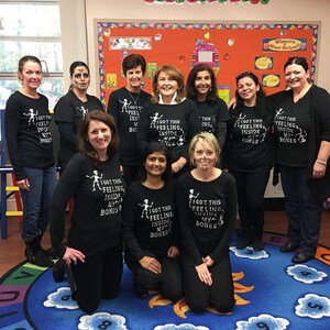 May include: A group of women wearing black t-shirts with a skeleton graphic and the text "I got this feeling inside my bones". The women are standing in a classroom with a blue and yellow rug on the floor.