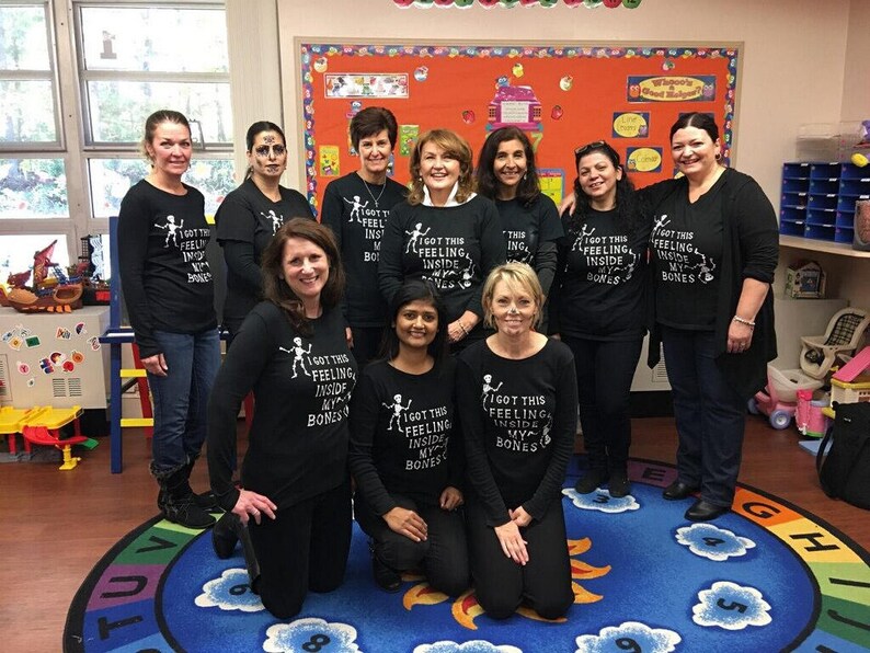 May include: A group of women wearing black t-shirts with a skeleton graphic and the text "I got this feeling inside my bones". The women are standing in a classroom with a blue and yellow rug on the floor.
