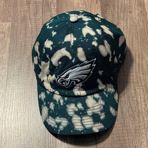 May include: A green and white tie-dye baseball cap featuring the Philadelphia Eagles logo. The cap has a curved brim and an adjustable strap. The Eagles logo is embroidered on the front in silver and white.