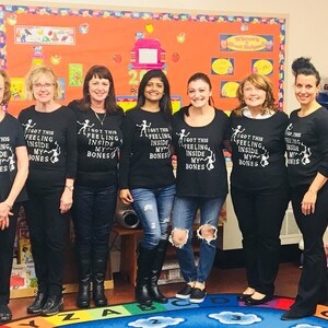 May include: Seven people wearing black t-shirts with the text "I got this feeling inside my bones" and a skeleton graphic. The group is standing in front of a colorful wall with a bulletin board.