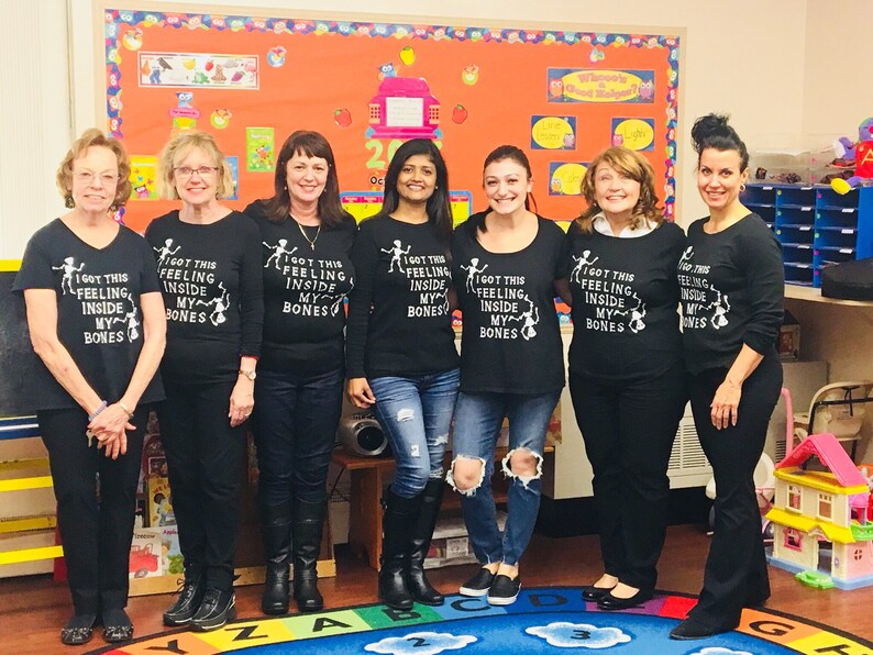 May include: Seven people wearing black t-shirts with the text "I got this feeling inside my bones" and a skeleton graphic. The group is standing in front of a colorful wall with a bulletin board.