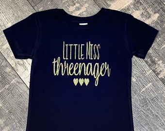 May include: A navy blue t-shirt with the text "Little Miss Three-nager" in gold glitter lettering and three gold glitter hearts.