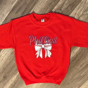 May include: A red crewneck sweatshirt featuring the word "Phillies" in script with a baseball-themed bow graphic. The design is in white and blue. The sweatshirt is made of a soft material.