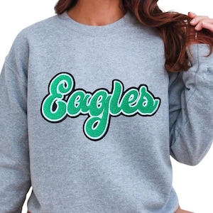 May include: A light gray sweatshirt with the word "Eagles" in a green, cursive font with a white and black outline. The sweatshirt has long sleeves and a crew neck.