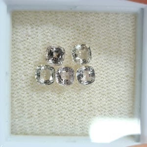 Könnte beinhalten: Fünf kissenförmige Edelsteine in einem weißen Display. Die Edelsteine haben eine blasse, neutrale Farbe mit subtilen Variationen. Das Gehäuse ist mit einem strukturierten, cremefarbenen Material ausgekleidet.