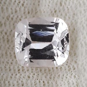 May include: A faceted, rectangular-shaped gemstone with a clear, colorless appearance. The gem has a slightly rounded shape and is cut to maximize light reflection, creating a brilliant sparkle. The gem is set against a white background.