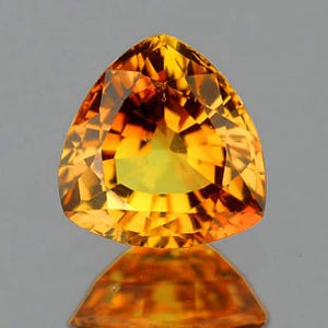 Op de afbeelding: Een gefacetteerde, driehoekige edelsteen met een briljante, goudgele tint. De ingewikkelde slijpsels van de edelsteen reflecteren het licht en creëren een oogverblindend schouwspel. De steen is geplaatst tegen een neutrale achtergrond, die de helderheid en kleur benadrukt. De reflectie van de edelsteen is hieronder zichtbaar.
