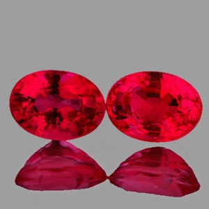 May include: Two oval-shaped, deep red gemstones with a faceted cut. The gemstones are set against a gray background.
