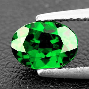 Puede incluir: Una gema ovalada de color verde vibrante con un corte facetado, sostenida en una pinza.