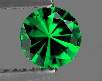 May include: A round, faceted, brilliant-cut emerald green gemstone held by a pair of silver tweezers. The gemstone's facets reflect light, creating a starburst pattern. The gem is a vibrant, saturated green color.