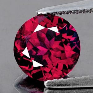 May include: A round, faceted, red gemstone with a deep, rich colour. The gemstone is held in a pair of tweezers.