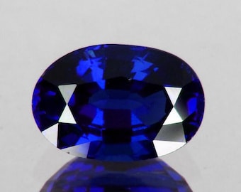Könnte beinhalten: Ein tiefblauer, oval geschliffener Edelstein mit reflektierenden Facetten. Der Edelstein ist vor einem weißen Hintergrund platziert, mit seiner Reflexion darunter. Der saphirähnliche Stein hat eine polierte Oberfläche und einen brillanten Glanz.