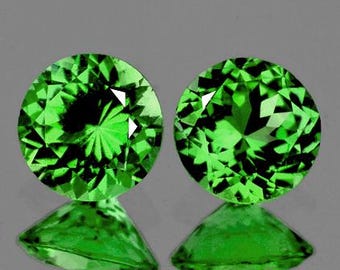 May include: Two round, faceted, bright green gemstones with a brilliant cut. The gems are displayed on a reflective surface, creating a mirrored effect. The stones are a vibrant, saturated green color.