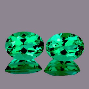 Op de afbeelding: Twee ovale groene edelstenen met een facetgeslepen vorm. De edelstenen zijn geplaatst tegen een grijze achtergrond.