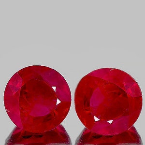 Puede incluir: Dos rubíes redondos y facetados. Las piedras preciosas son de un color rojo intenso y vibrante, con facetas que capturan la luz. Los rubíes se muestran sobre un fondo neutro, resaltando su color y brillo.
