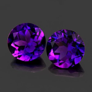 Puede incluir: Dos gemas de amatista redondas y facetadas. Las gemas muestran un tono púrpura intenso con destellos de violeta y azul más claros. Las piedras están sobre una superficie oscura y reflectante.