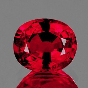 Puede incluir: Una gema facetada, de corte ovalado, con un tono rojo intenso. La gema tiene una superficie reflectante, con facetas triangulares y rectangulares que crean un juego de luces y sombras. Sobre un fondo gris.