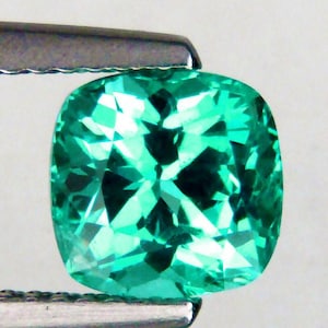 Op de afbeelding: Een vierkant geslepen, levendige groene edelsteen, vastgehouden door een metalen gereedschap. De edelsteen heeft een gefacetteerd oppervlak dat licht reflecteert, waardoor een sprankelend effect ontstaat. De kleur van de edelsteen is een rijk, verzadigd groen en de slijpvorm is symmetrisch.