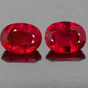 May include: Two oval-shaped, deep red gemstones with a polished finish. The gemstones are set against a gray background.