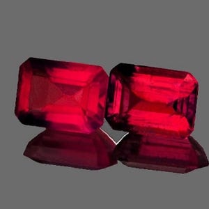 May include: Two radiant, rectangular-cut, deep red gemstones. The gems are highly polished, reflecting light and creating a brilliant shine. The stones are set against a gray background, highlighting their color and clarity. These gems are likely for jewelry.